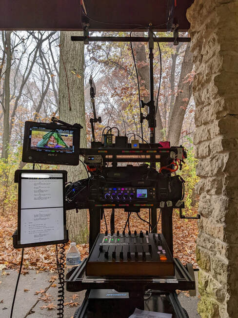 A feature film sound cart with recorder and faders, along with a monitor, script, and antennas, is on an exterior forest set