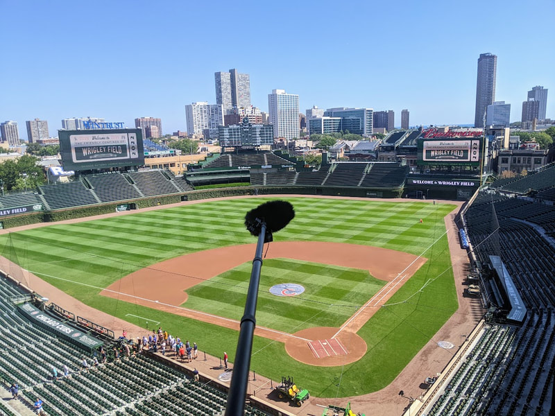 A boom pole with microphone extends into the air over Wrigley Field in Chicago, IL with blue skies and tall buildings in the background